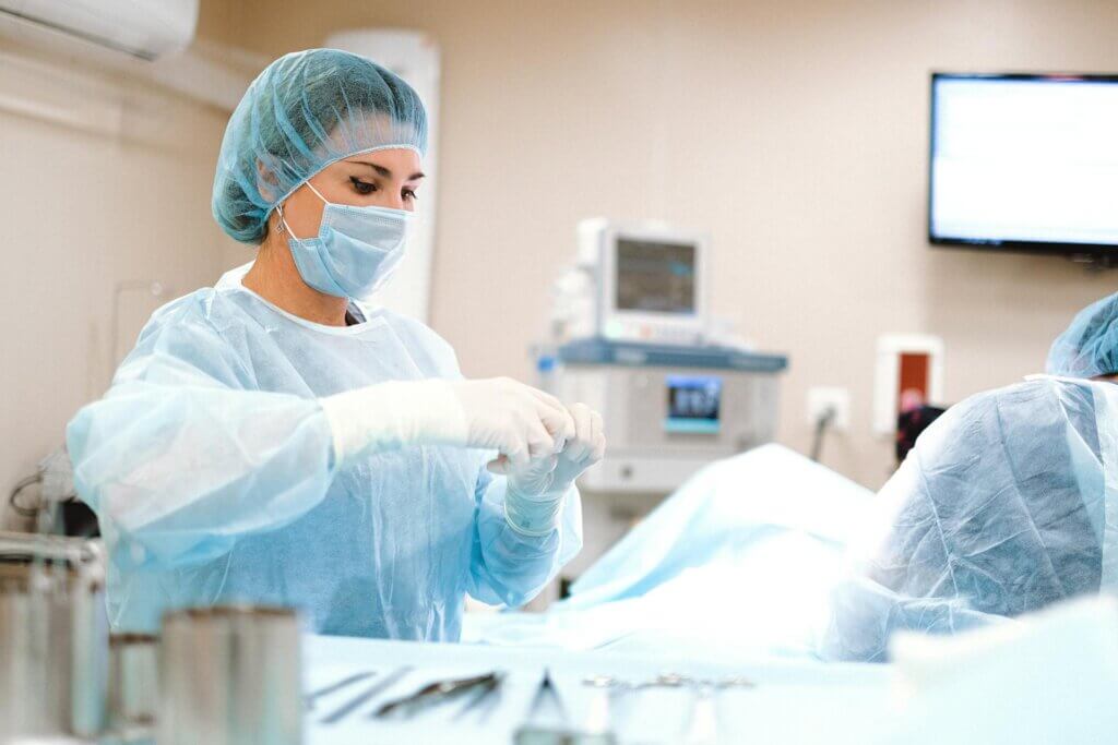 A surgeon wearing a blue surgical gown, cap, face mask, and gloves preparing equipment in a hospital operating room.
