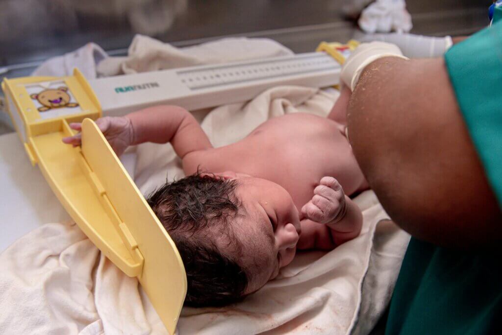 A newborn infant lying on a medical scale being measured by a healthcare provider in a hospital delivery room.