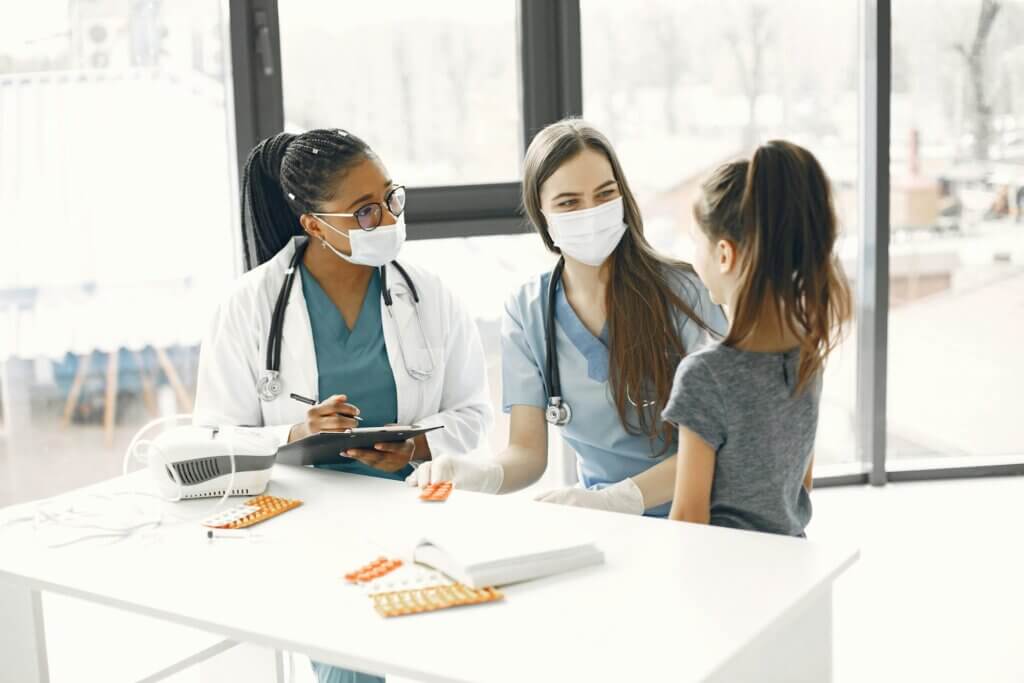 Two medical professionals wearing masks and scrubs talking with a young girl patient in a clinic room.