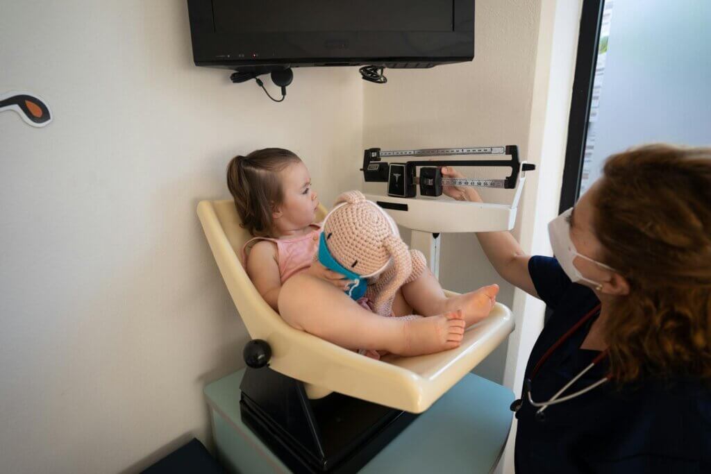A nurse wearing a mask and scrubs measuring the weight of a young child holding a stuffed toy on a scale in a clinic room.