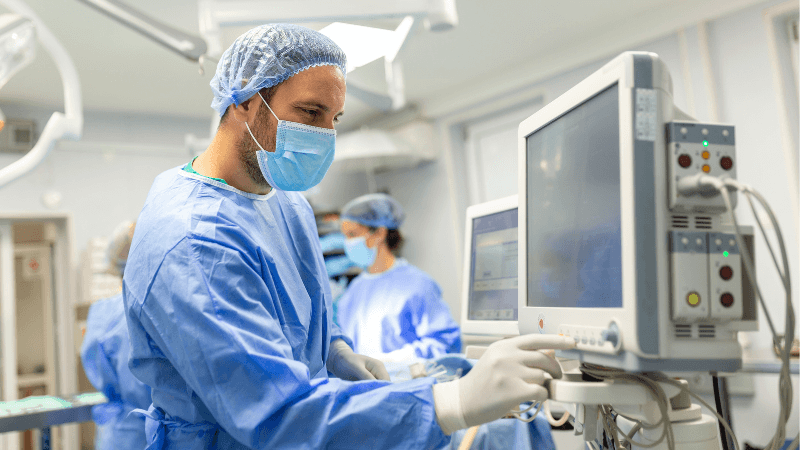 Surgeon wearing light blue scrubs, a surgical cap, a face mask, and gloves adjusting a medical monitor or equipment in an operating room.