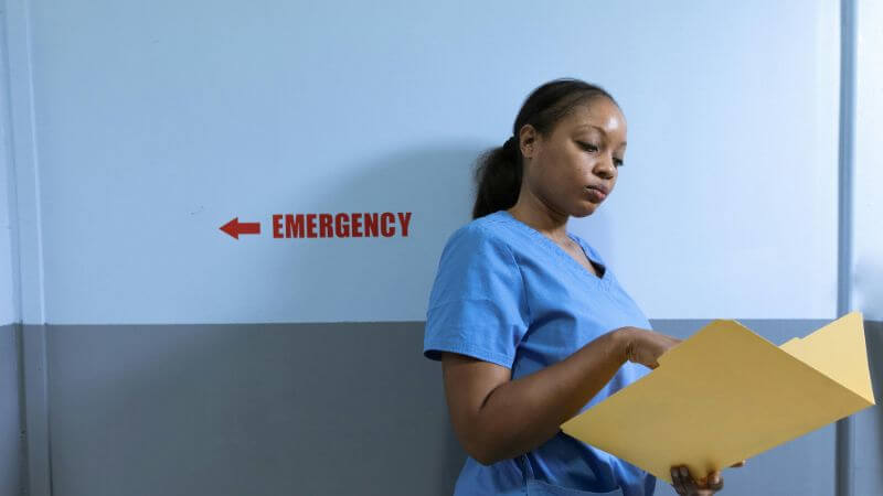 A healthcare professional in blue scrubs reviewing a yellow folder while standing in a hospital hallway next to an "Emergency" sign with an arrow.