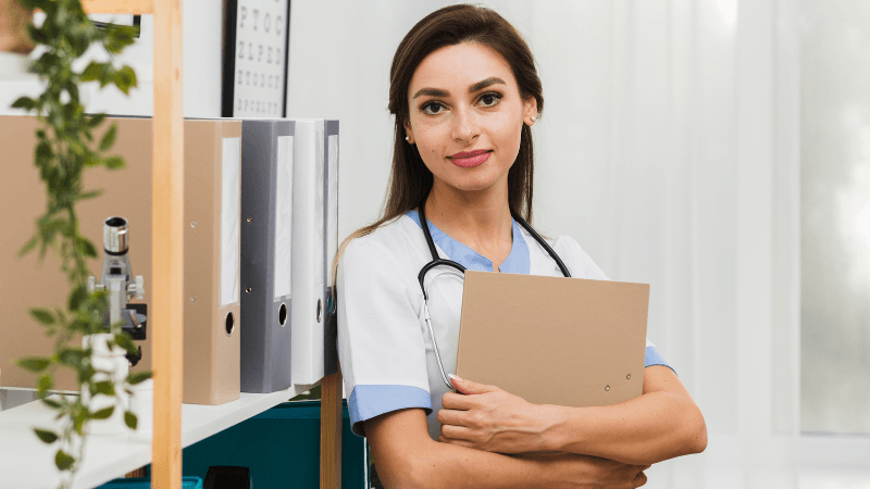 A smiling female healthcare professional or nurse wearing white and blue scrubs with a stethoscope, holding a folder near a bookshelf with binders in an office.