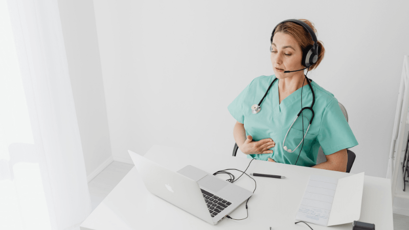 A female healthcare professional or nurse wearing turquoise scrubs, a stethoscope, and a headset sitting at a desk with a laptop and documents.