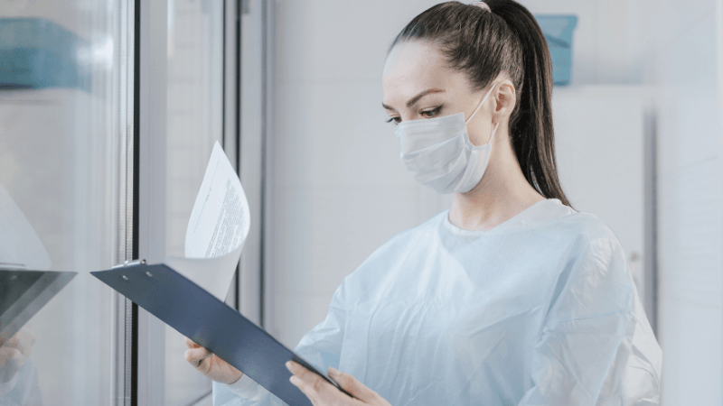 A female doctor or healthcare professional wearing a face mask and medical gown examining patient medical records on a clipboard.