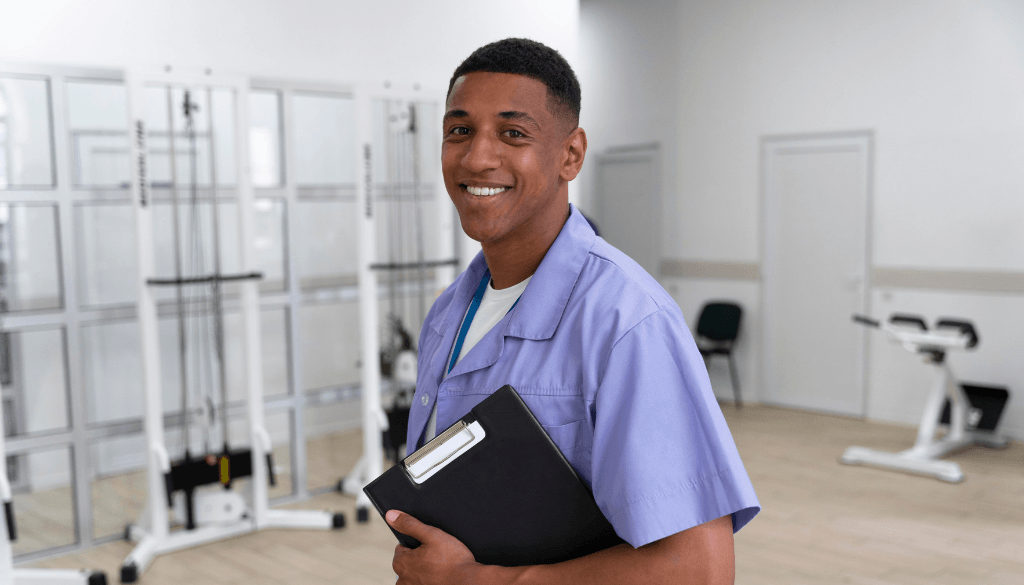 A smiling Ghanaian healthcare professional in light purple scrubs holding a black clipboard inside a medical facility or rehabilitation center with exercise equipment.