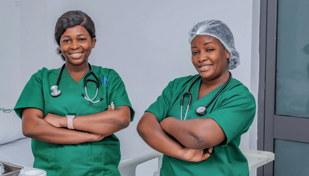 Two smiling female Kenyan healthcare workers wearing green medical scrubs with stethoscopes around their necks and arms crossed in a hospital room.