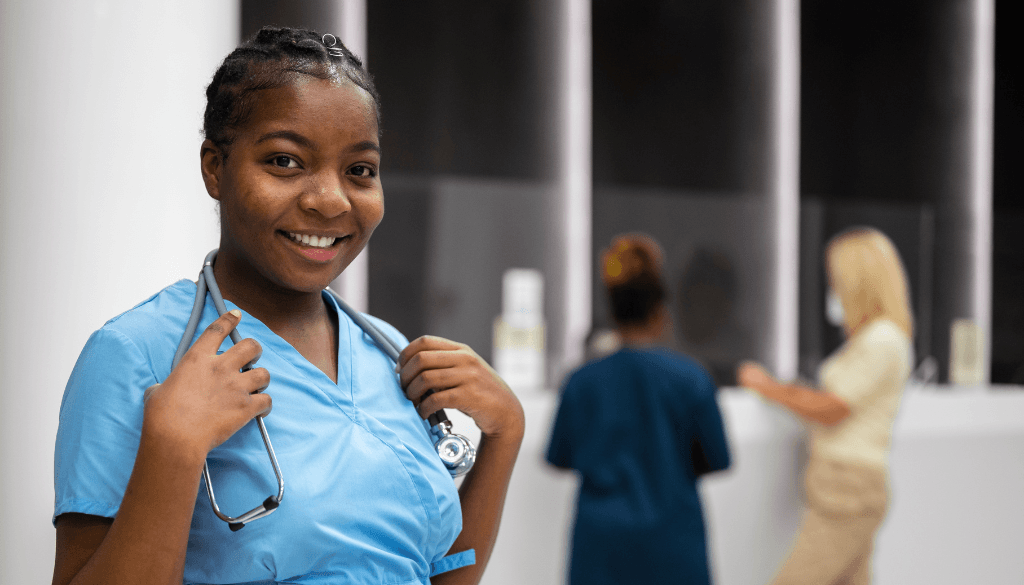 A smiling Nigerian nurse wearing blue scrubs and holding a stethoscope around her neck inside a hospital corridor, with other medical staff in the background.