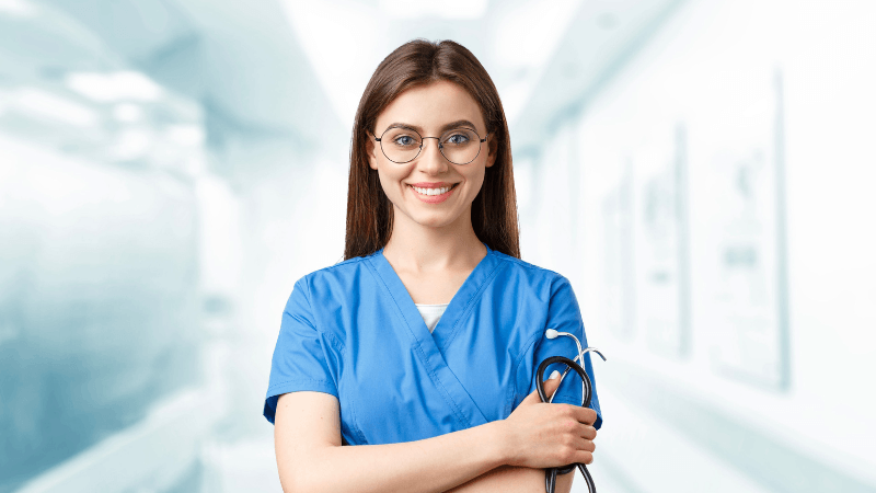 A smiling female nurse wearing blue scrubs, round glasses, and a stethoscope with arms crossed, standing in a bright hospital hallway.