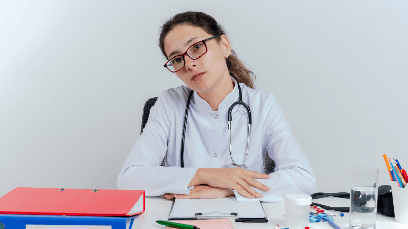 A female doctor wearing glasses, a white lab coat, and a stethoscope sitting at a desk with medical files, a container, and pills.