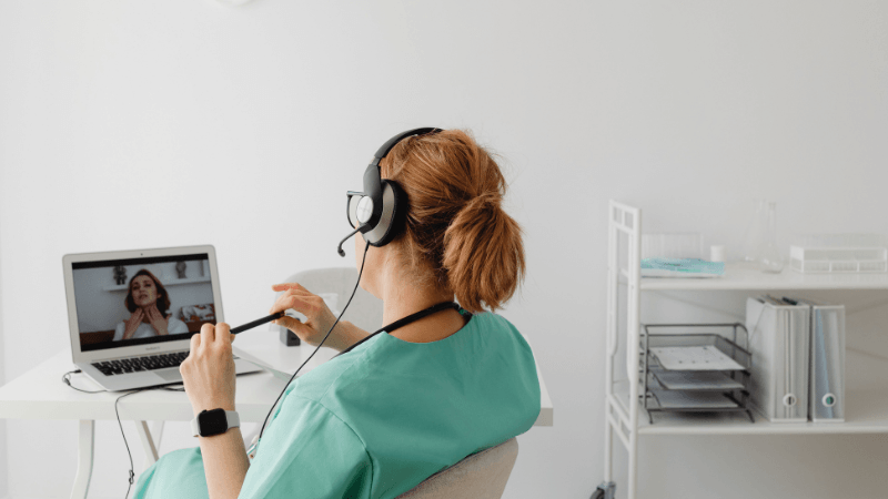 A nurse wearing green scrubs, a stethoscope, and a headset sitting at a desk and participating in a video call on a laptop.