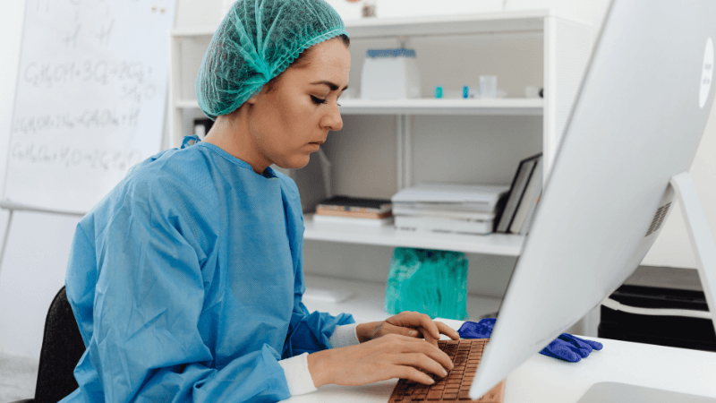 A nurse wearing blue scrubs and a surgical cap typing on a keyboard at a desk in front of a computer monitor in an office.