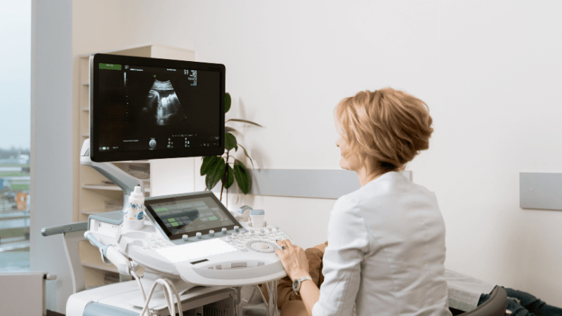 A female medical professional or sonographer operating an ultrasound machine with a scan displayed on the monitor in a clinic room.