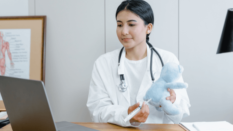 A female healthcare professional or nurse wearing a white coat and stethoscope sitting at a desk with a laptop while demonstrating an injection technique on a blue stuffed toy.