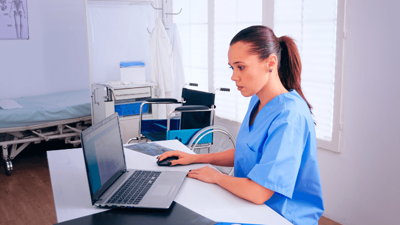 A female nurse wearing blue scrubs sitting at a desk and working on a laptop in a hospital room with a wheelchair and medical bed in the background.