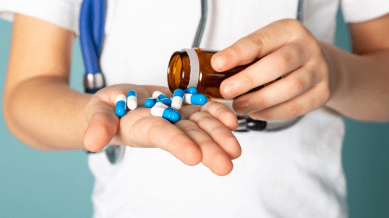Healthcare professional wearing a stethoscope and white shirt pouring blue and white capsules from a prescription medicine bottle into their hand.
