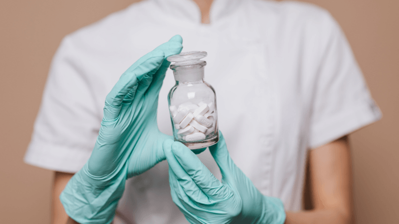 Healthcare professional wearing mint green medical gloves and a white uniform holding a glass bottle filled with white pills.