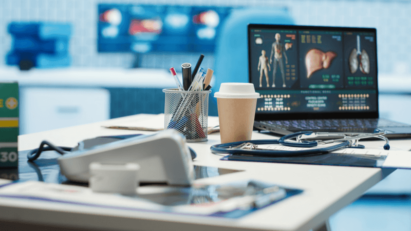 Medical office desk featuring a laptop displaying human anatomy charts and internal organs, a stethoscope, a coffee cup, a pen holder, and medical equipment.