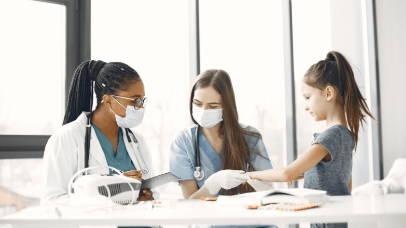 Two female healthcare professionals wearing masks and stethoscopes examining a young female patient in a bright clinic setting.