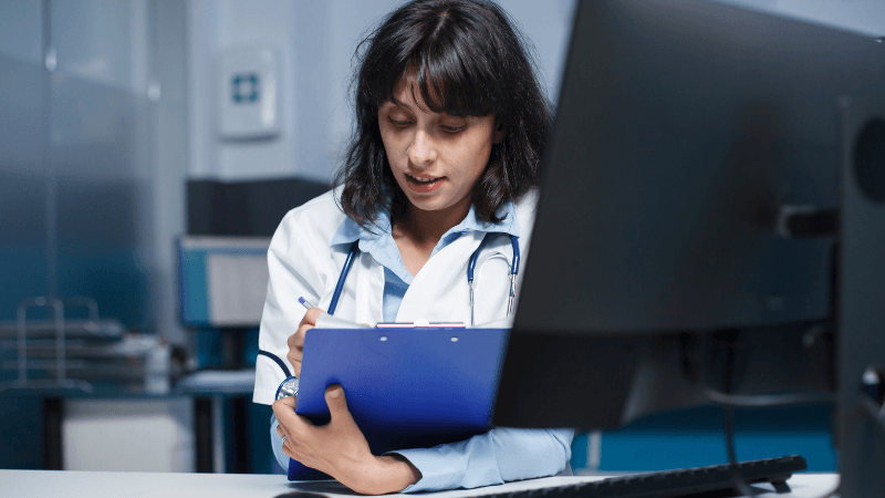 A female doctor wearing a white lab coat and stethoscope writing notes on a blue clipboard while sitting at a desk next to a computer monitor.