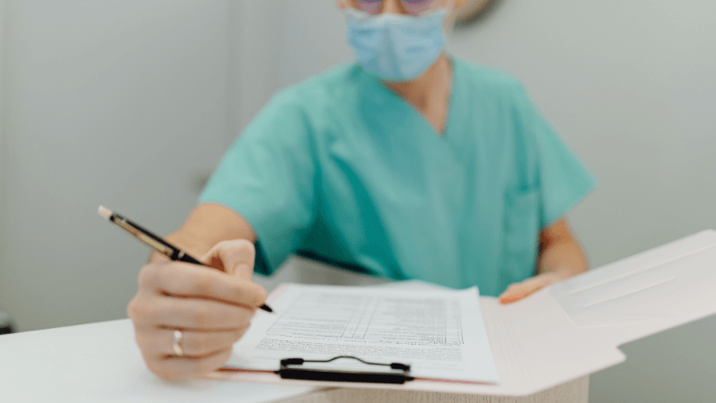 A healthcare worker wearing turquoise scrubs and a face mask writing notes on a document clipboard with a pen.