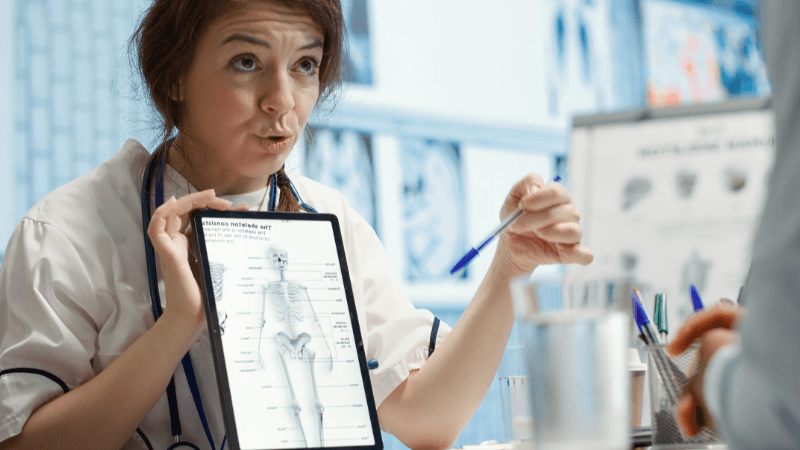 A female doctor wearing a white coat and stethoscope holding up a tablet displaying a human skeletal anatomy chart while explaining details to a colleague in an office.