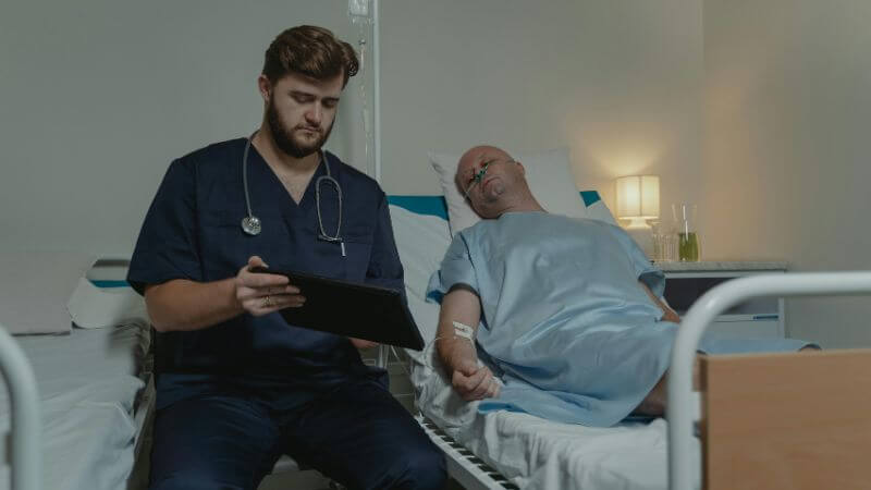 A healthcare professional in dark blue scrubs with a stethoscope checking a digital tablet while sitting beside a patient resting in a hospital bed.