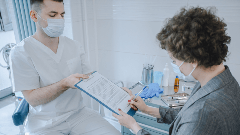 A male healthcare worker wearing a white uniform and face mask handing a clipboard to a female patient who is signing a medical document with a pen.