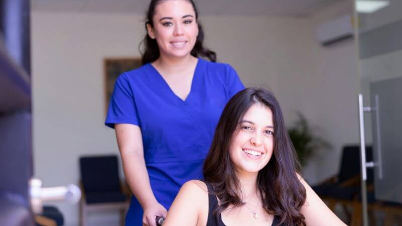 A nurse in blue scrubs assists a smiling patient.