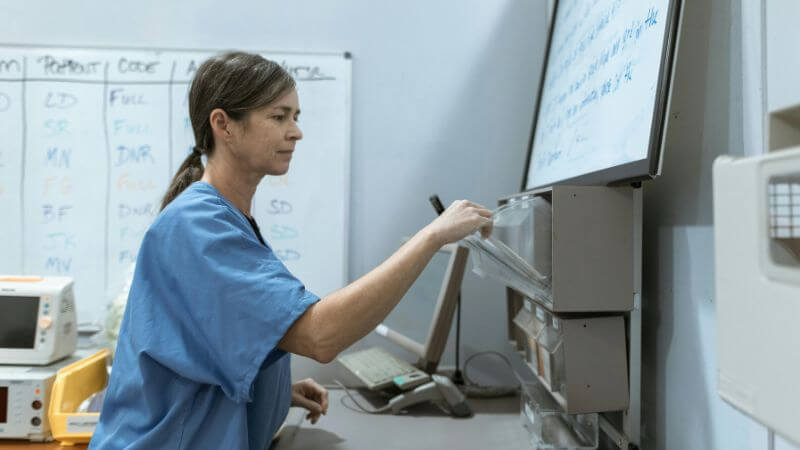 A healthcare professional in blue scrubs organizing files or supplies at a hospital station with a whiteboard and chart bins in the background.