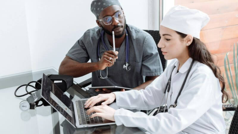 Two medical professionals working together, with a female healthcare worker typing on a laptop and a male doctor in dark scrubs and glasses holding a tablet and stethoscope.