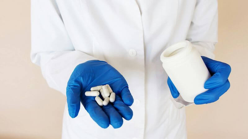 Healthcare worker in blue gloves holding capsules and a medicine bottle.