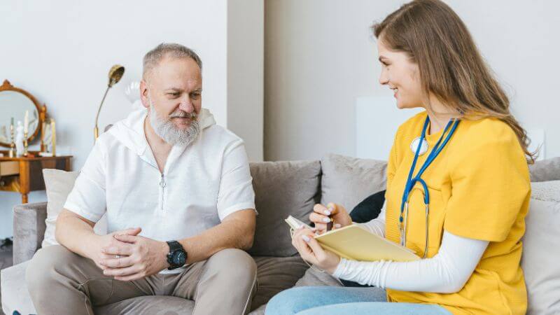 A smiling healthcare professional in a yellow top and stethoscope taking notes in a notebook while consulting with an older male patient sitting on a couch.