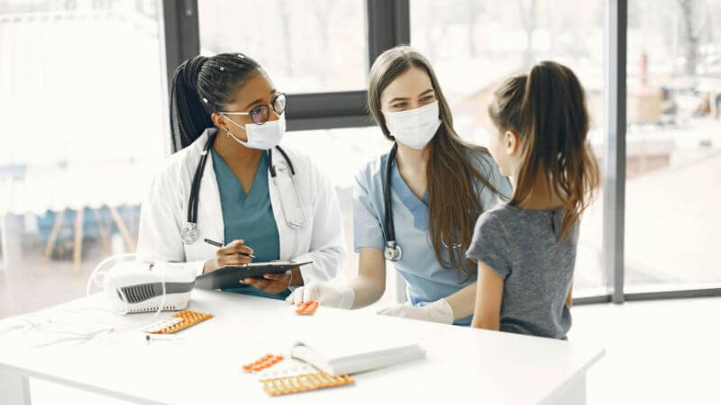 Two female doctors wearing face masks and stethoscopes consulting with a young girl patient across a table with medical supplies and blister packs.