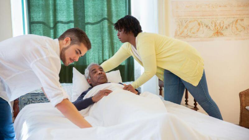 Two caregivers helping an elderly man lying comfortably in a bed with white sheets and pillows.