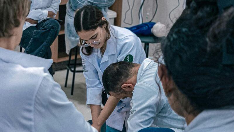 Medical students and instructors in white coats practicing hands-on clinical procedures and first aid techniques during a training workshop.