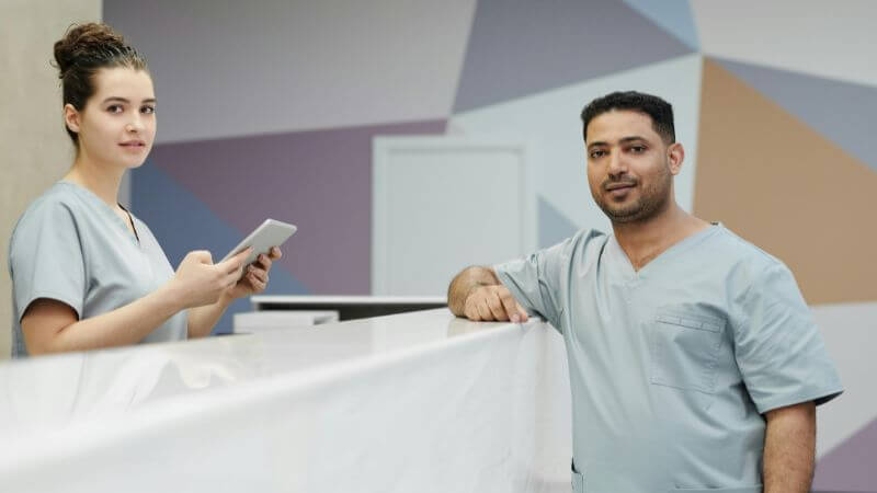Two healthcare professionals in light gray scrubs standing behind a clean reception desk, with a female nurse holding a tablet and a male medical staff member leaning against the counter.
