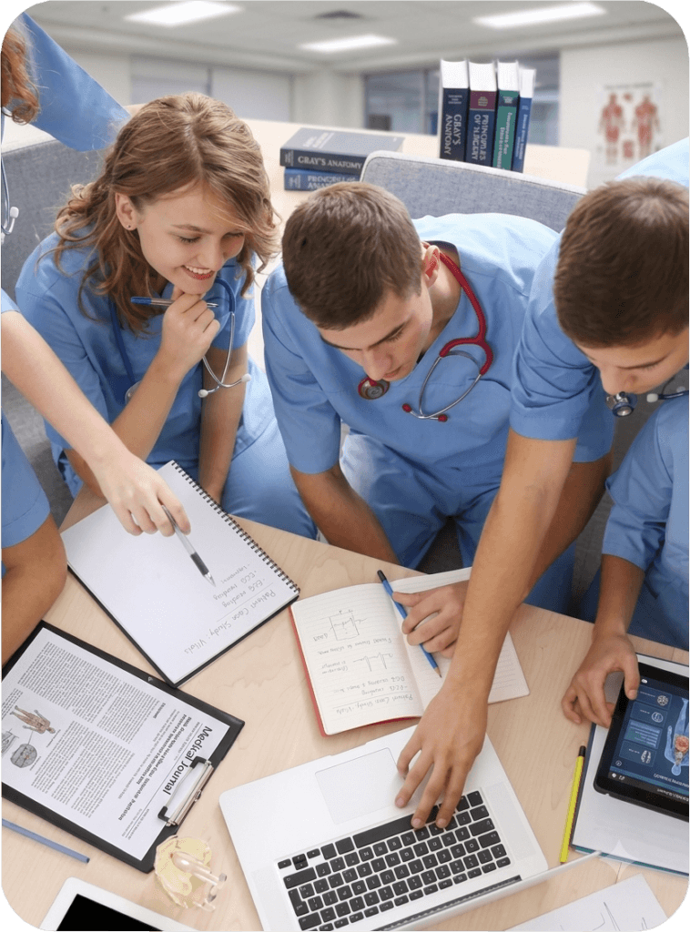 Group of nursing students and professionals wearing blue scrubs collaborating around a table, reviewing medical journals, writing in notebooks, and using a laptop.