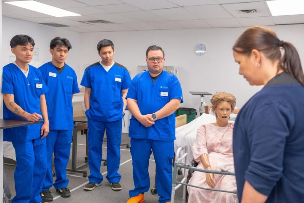 Nursing students in blue scrubs practicing patient care using a medical mannequin.