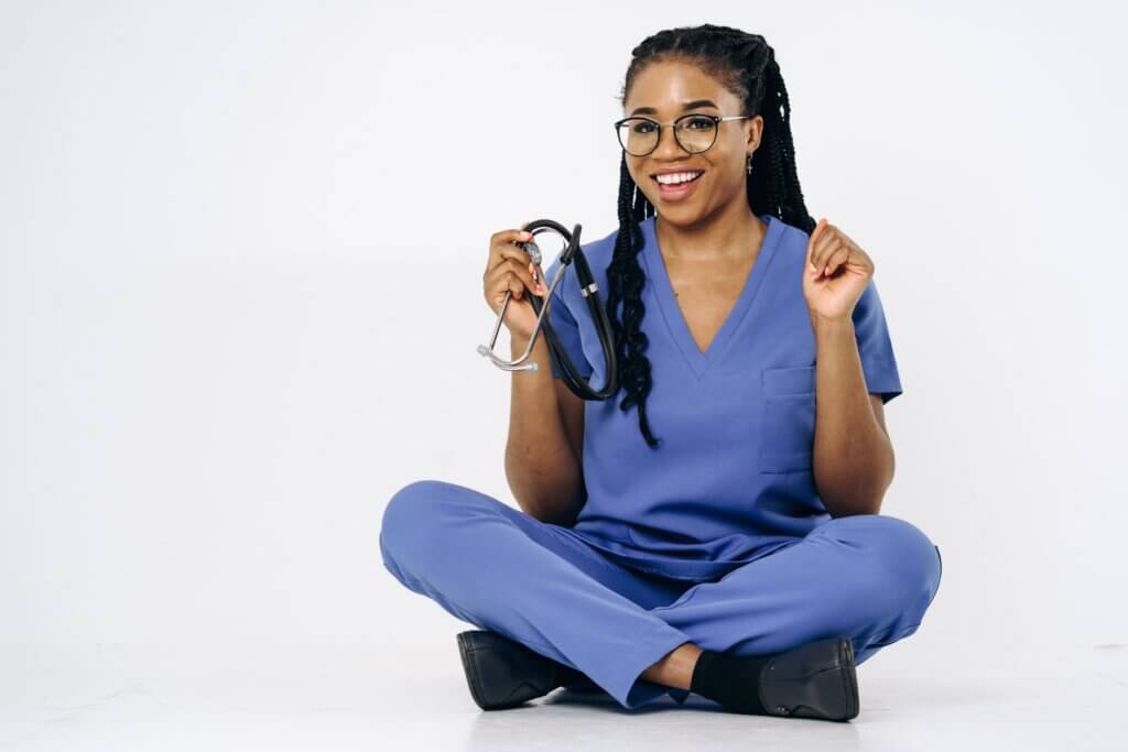 A cheerful nurse in blue scrubs holding a stethoscope while sitting
